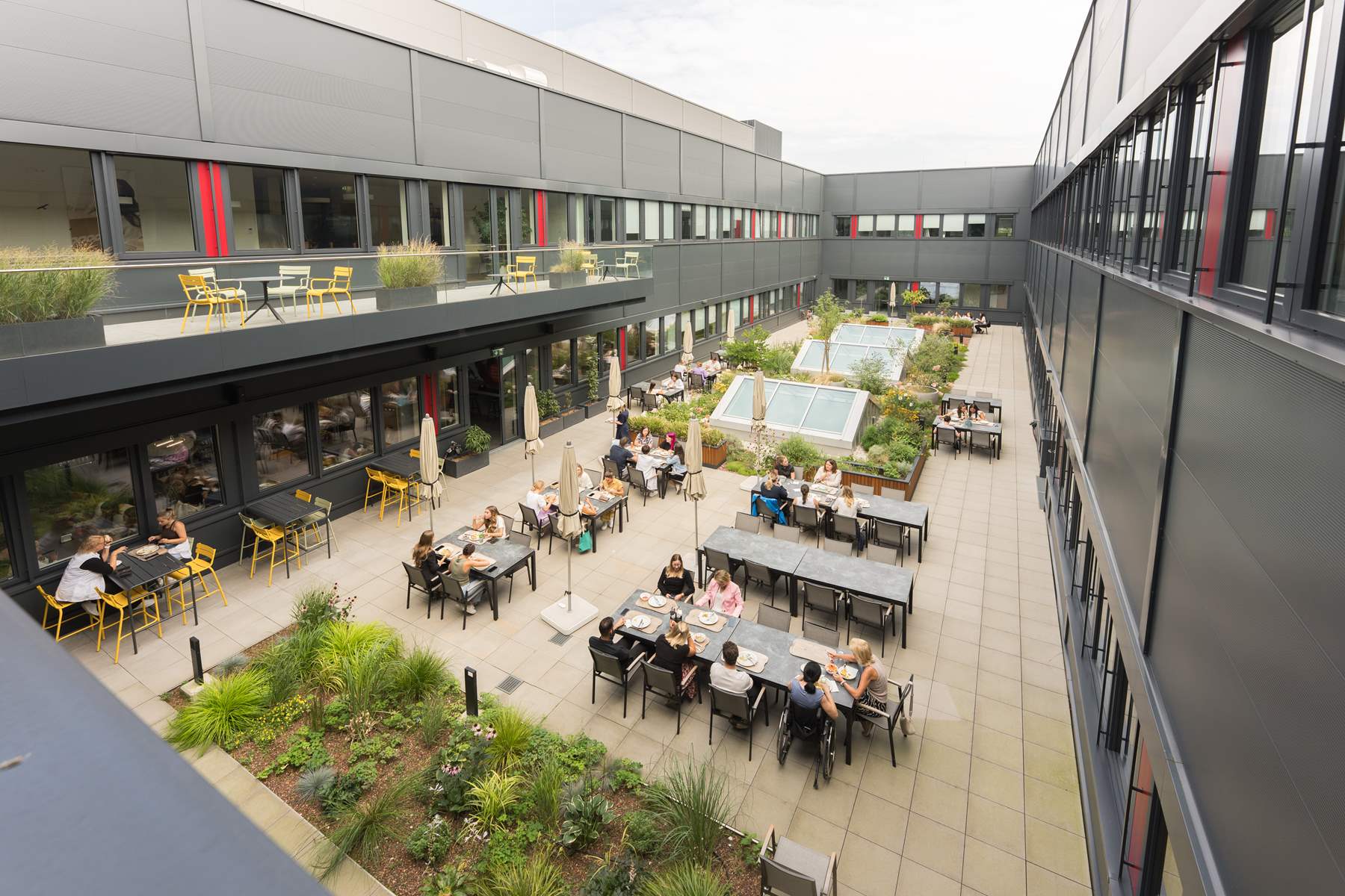 In einem Innenhof eines Bürogebäudes stehen mehrere Bänke und Tische auf denen Menschen gerade zu Mittag essen.